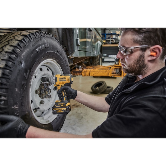 A person wearing gloves uses a DEWALT cordless impact wrench (model DCF891N) to fasten or remove bolts from a vehicle wheel in a workshop environment. The person's face is blurred for privacy.