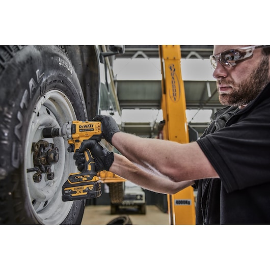 A person wearing black gloves uses a DEWALT DCF891P2G cordless impact wrench to tighten bolts on a vehicle wheel in a workshop setting.