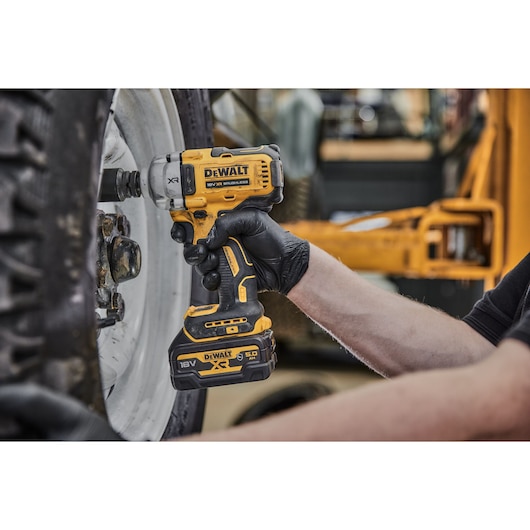A person wearing black gloves is using a yellow DEWALT cordless impact wrench (model DCF891P2G, 18V XR) to work on the bolts of a large vehicle wheel.