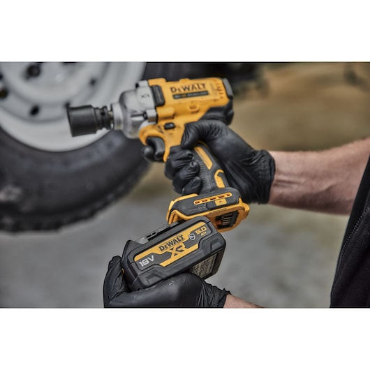 A person wearing black gloves is holding a yellow and black DEWALT cordless impact wrench and a DEWALT 18V XR 5.0Ah lithium-ion battery. The background shows a blurred tire.
