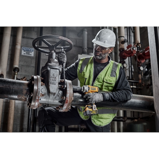 Worker uses DEWALT tools DCF892N, DCZ407P1T1-XE on industrial pipe in factory setting. Safety gear worn.