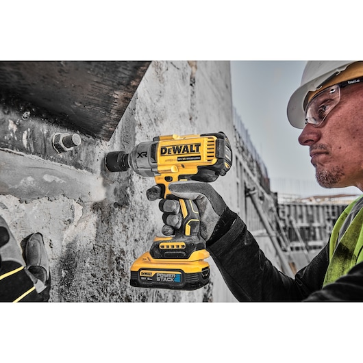 A person wearing work gloves uses a DEWALT cordless impact wrench (model DCF900H2T) with POWERSTACK battery to fasten a bolt into a concrete wall at a construction site.