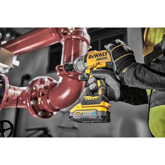 A person wearing work gloves operates a DEWALT cordless impact wrench (DCF900H2T) with a POWERSTACK battery, working on large industrial red pipes.