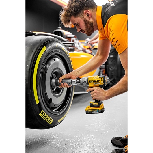 A person uses a DEWALT DCF900H2T cordless impact wrench to work on the wheel of a racing car. The scene takes place in a garage, with the racing car's Pirelli tire and details of the yellow DEWALT tool clearly visible.