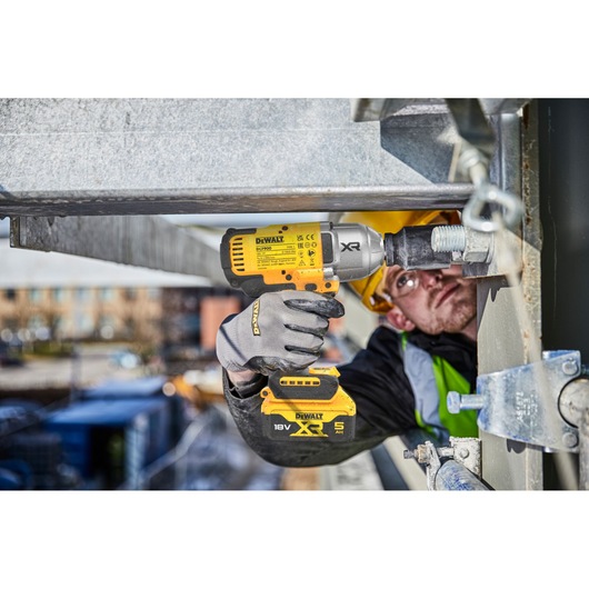 A person using a DEWALT DCF900NT-XJ cordless impact wrench to tighten a large bolt on a metal structure at a construction site. The person is wearing gloves, a safety vest, and a helmet.