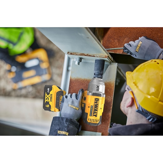 Construction worker using a yellow and black DEWALT DCF900N cordless impact wrench to fasten a large metal beam, wearing safety gloves and a yellow hard hat.