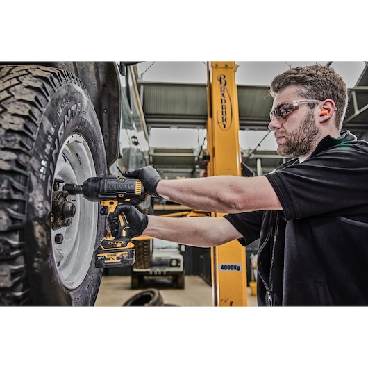 A person wearing gloves uses a DEWALT cordless impact wrench to remove or tighten lug nuts on a vehicle tire in an automotive workshop. The person's face is blurred, and there is workshop equipment and a lifted vehicle visible in the background.