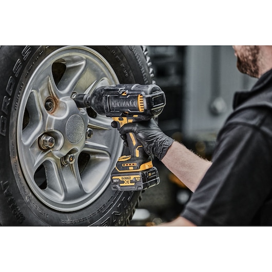 A person using a DEWALT cordless impact wrench to tighten or loosen lug nuts on a car wheel. The person is wearing a black glove, and the DEWALT power tool features yellow and black colors.