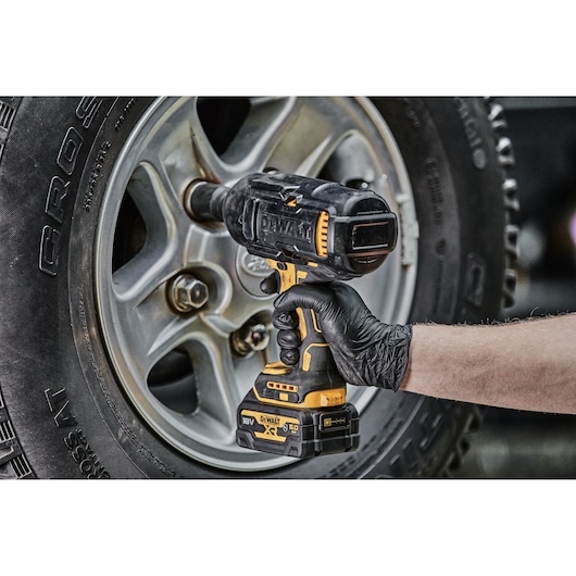 A hand wearing a black glove uses a DEWALT cordless impact wrench to tighten or loosen lug nuts on a car wheel.