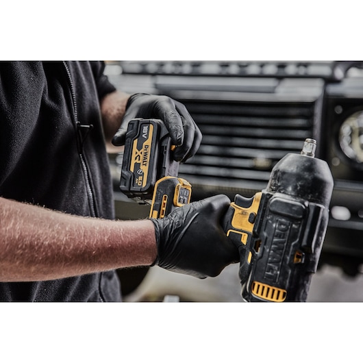 Close-up of a person wearing black gloves installing a DEWALT 18V XR battery into a cordless power tool. The tools and battery are black and yellow, and the background shows part of a vehicle grille.