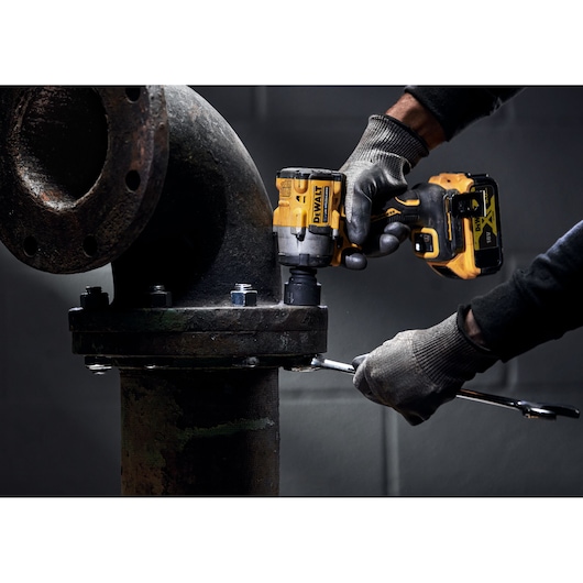 A person wearing gloves uses a DEWALT cordless impact wrench to tighten bolts on a large industrial pipe.