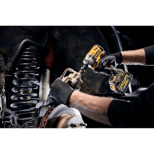 A person wearing black gloves uses a DEWALT cordless impact wrench to work on a vehicle's suspension and brake components.