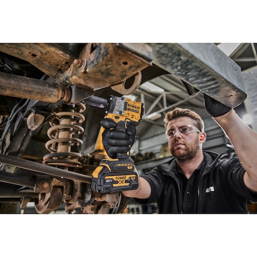 A person wearing black gloves uses a DEWALT 18V XR cordless impact wrench (model DCF921P2G) to work on a vehicle's suspension system in a workshop. The person's face is blurred.