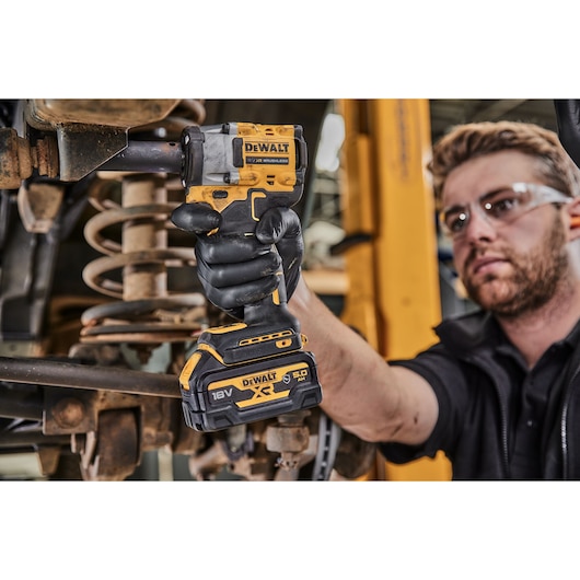 A person wearing black gloves uses a DEWALT 18V cordless impact wrench to work on machinery. The person's face is blurred, and the tool is clearly visible in the foreground.