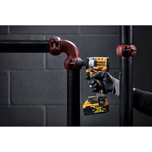 A person wearing black gloves uses a DEWALT cordless impact wrench to tighten a bolt on a metal pipe joint. The tool is yellow and black, and the battery pack is visible. The background features a grey brick wall and industrial piping.