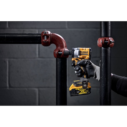 A person wearing a black and grey work glove is using a DEWALT 18V cordless impact wrench on a black metal pipe fitting in an industrial setting. The background is a grey brick wall.