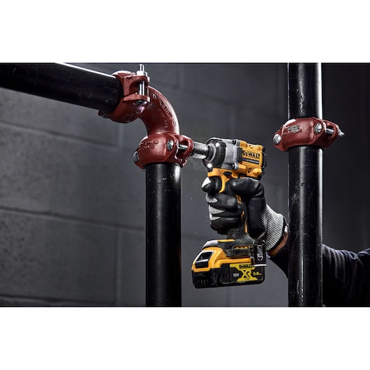 A person wearing work gloves uses a yellow and black DEWALT cordless impact wrench to tighten a bolt on a black metal pipe with a red fitting.