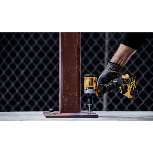 A person wearing a glove is using a DEWALT cordless impact wrench to tighten a bolt on a metal base plate attached to a vertical steel beam.