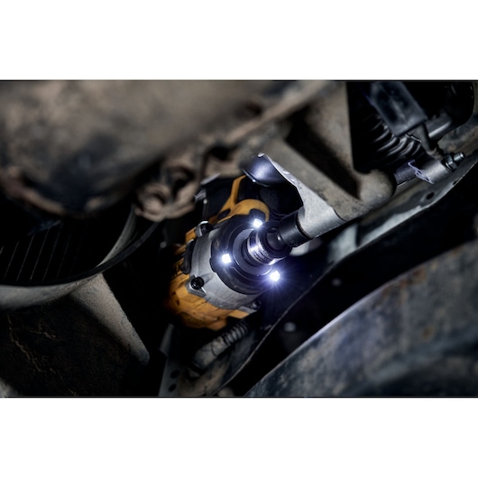 A close-up view of a DEWALT power tool with LED lights being used to work on a bolt underneath a vehicle, illuminating a dark engine or machinery area.