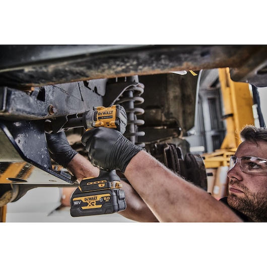 A person wearing black gloves uses a DEWALT 18V XR cordless impact wrench to work on the undercarriage of a vehicle in a workshop.