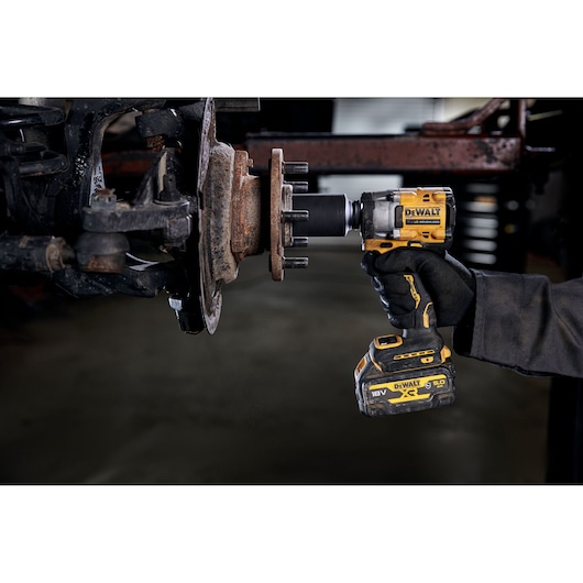 A person using a DEWALT cordless impact wrench (model DCF923P2G) to work on a rusty vehicle wheel hub in a garage or workshop.