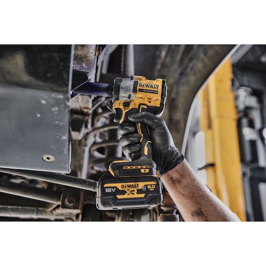 A person wearing black gloves uses a DEWALT DCF923P2G 18V cordless impact wrench to work on machinery or a vehicle part.