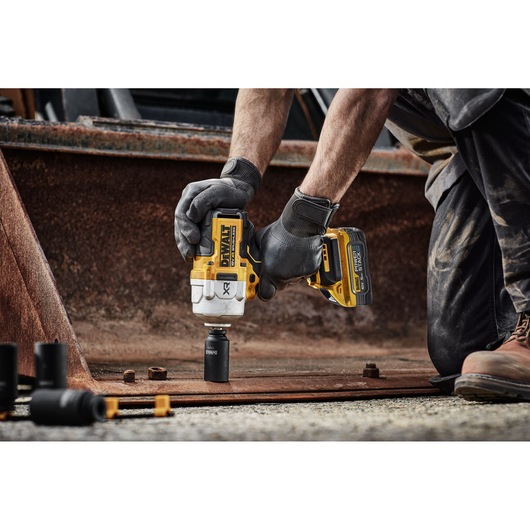A person wearing work gloves uses a yellow and black DEWALT cordless impact wrench to loosen a bolt on a rusty metal surface at an industrial site.
