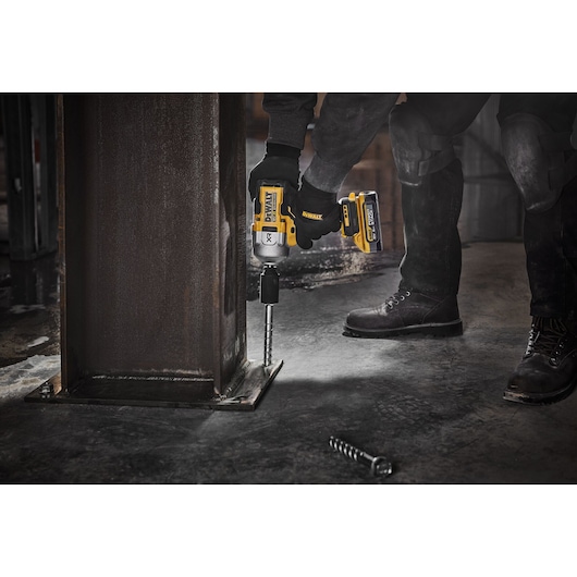 A person wearing gloves and work boots uses a DEWALT cordless power tool to fasten a bolt to a metal structure in an industrial setting.