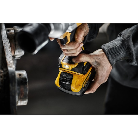 Close-up of a person holding and operating a yellow and black DEWALT cordless power tool, with visible wear and dirt on their hands. The tool appears to be used for working on metal parts.