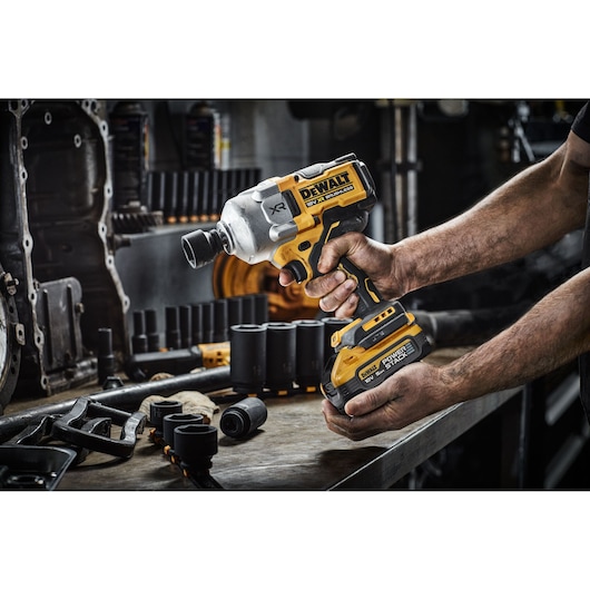 A person holds a DEWALT cordless impact wrench, model DCF961H2T, with a battery attached. The tool is being used in a workshop with various sockets and mechanical parts on the workbench.