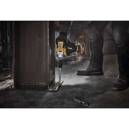 A person using a DEWALT cordless impact wrench to fasten a large bolt into the base of a metal beam in an industrial setting. Another person stands nearby, both wearing work boots and protective knee pads.