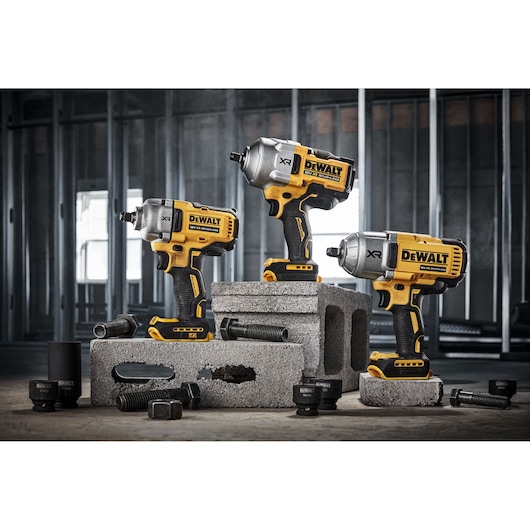 Three DEWALT cordless impact wrenches are displayed on concrete blocks in a construction setting, surrounded by large bolts and sockets.