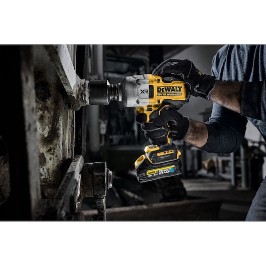 A close-up of a person using a DEWALT cordless impact wrench with a POWERSTACK battery to work on industrial machinery. The person is wearing black gloves and a long-sleeved shirt.