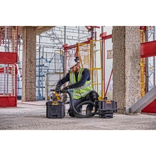 A construction worker wearing a white helmet, gloves, and a reflective safety vest is using a DEWALT power tool connected to dust extraction equipment on a construction site. The worker is kneeling on the ground surrounded by concrete columns and scaffolding.