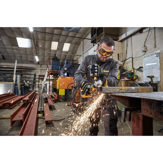 A person using a DEWALT portable grinder to cut metal, with sparks flying in an industrial workshop setting. The person's face is blurred for privacy.
