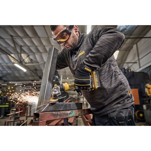 A person wearing protective gloves uses a DEWALT DCG404NT cordless angle grinder to cut metal, producing sparks, in an industrial workshop setting. The person's face is obscured.