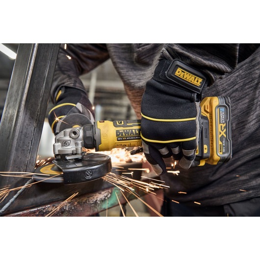 A close-up of a person wearing gloves and operating a DEWALT cordless angle grinder, creating sparks while grinding a metal surface.
