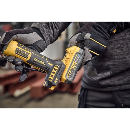 Close-up of a person wearing protective gloves holding a yellow DEWALT cordless tool, specifically an angle grinder with a brushless motor and XR battery, in an industrial setting.