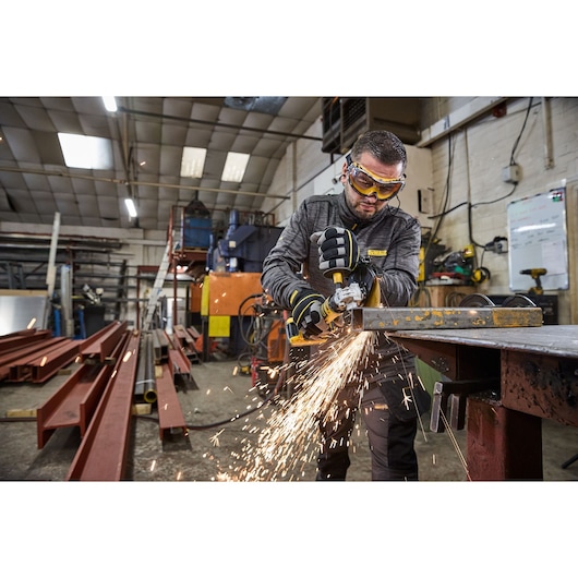 A person using a DEWALT cordless angle grinder to cut a metal beam in an industrial workshop. Sparks are flying from the metal as they work. The person is wearing gloves and protective clothing.