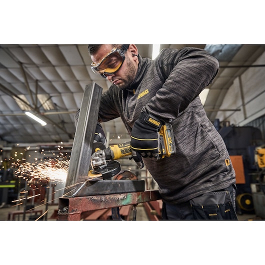 A person wearing safety gloves is using a DEWALT cordless angle grinder on a metal beam, generating sparks, inside an industrial workshop.