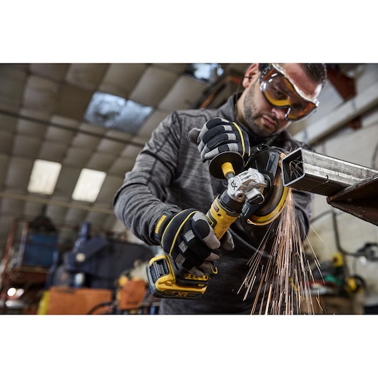 A person wearing safety gloves is using a DEWALT cordless angle grinder to cut a metal piece, producing sparks, in an industrial workshop setting. The person's face is blurred for privacy.