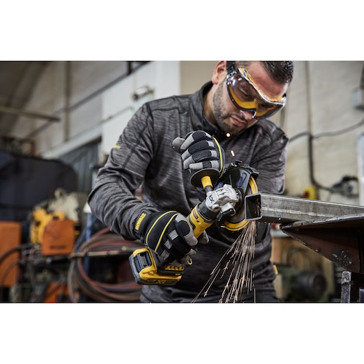A person wearing protective gloves is using a DEWALT cordless angle grinder (model DCG404N) to cut a metal piece, with sparks flying in an industrial workshop setting.