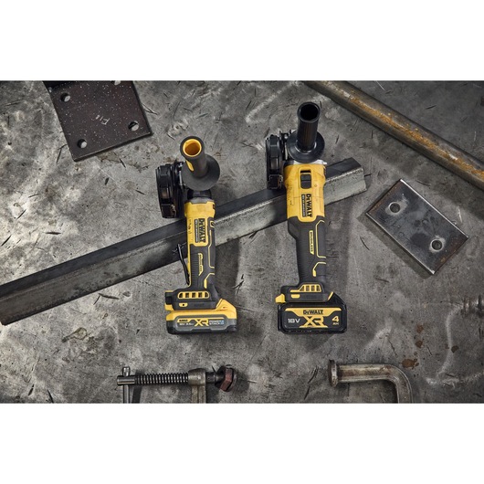 Two DEWALT cordless angle grinders, both labeled XR 18V, are placed side by side on a metal workbench. Metal bars, plates, and clamps are scattered around them.