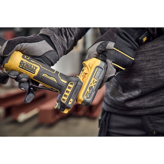 Close-up of a person wearing gloves holding a DEWALT 18V XR brushless cordless power tool with a battery attached, shown in a workshop setting.