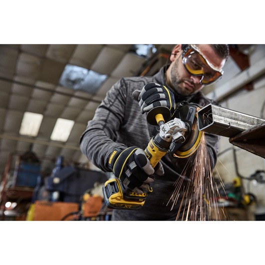 A person wearing protective gloves uses a yellow DEWALT cordless angle grinder to cut through a metal pipe in an industrial workshop, with sparks flying from the cutting area.