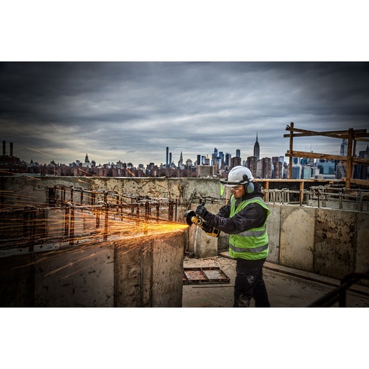 A person wearing gloves using a DEWALT cordless angle grinder to cut or grind concrete, with visible dust rising from the work area.