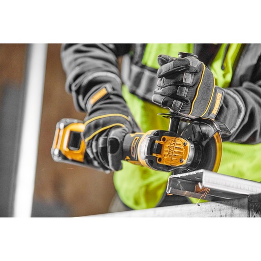 A person wearing protective gloves and a high-visibility vest is using a DEWALT cordless angle grinder to cut a metal piece.