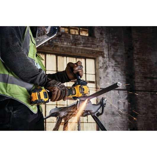 A person wearing protective gloves and a face shield uses a DEWALT cordless angle grinder to cut through a metal bar, with sparks flying, in an industrial workshop setting.