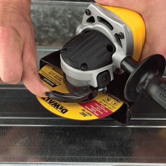 Close-up of a person adjusting the safety guard of a DEWALT angle grinder with a yellow body. The grinder is positioned on a metal surface.