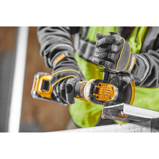 A person wearing safety gloves and a hi-vis vest using a DEWALT cordless angle grinder to cut a metal piece.